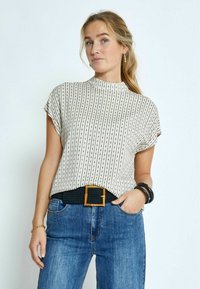 Kurzarmbluse in Creme mit schwarzen vertikalen Streifen, ausgestattet mit einem hohen Kragen. Kombiniert mit blauen Jeans und einem schwarzen geflochtenen Gürtel mit einer quadratischen Schnalle.