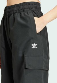 Svarta träningsbyxor med elastisk midja, sidofickor och en cargoficka. Har en vit Adidas-logotyp på vänster ben. Slät textur.