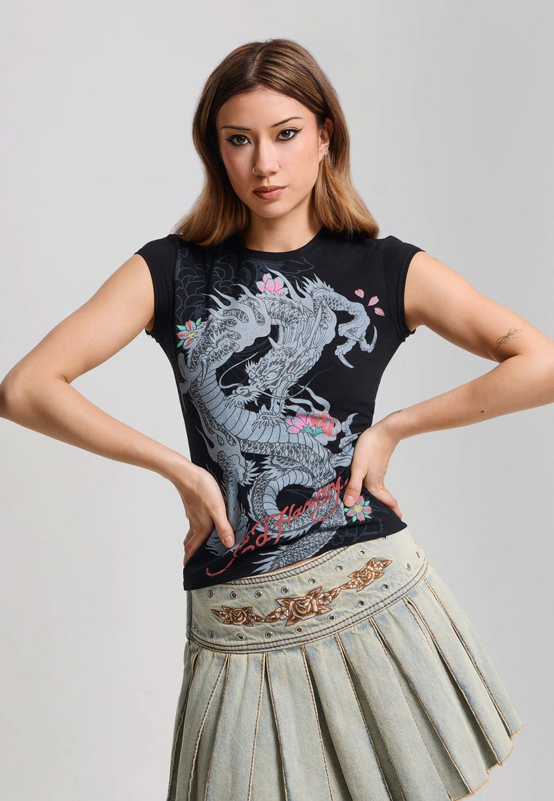 Jeune femme aux cheveux bruns et raides porte un T-shirt noir à motif de dragon et une jupe plissée en jean avec broderies florales, posant les mains sur les hanches.