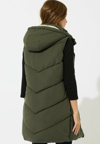 Olivengrüne gepolsterte Weste mit Kapuze, die ein Zickzack-Steppmuster aufweist und ein weiches Innenfutter hat. Ärmellos mit Seitentaschen.