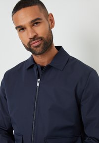 Marineblauer Zip-Jacke mit Kragen, hergestellt aus glattem Stoff. Verfügt über frontale Taschen und silberne Reißverschlussdetails. Minimalistisches Design.