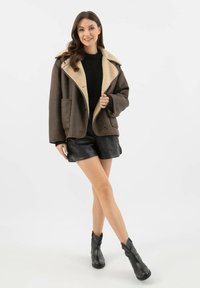 Hnědá umělohedvábná bunda s širokým límcem, knoflíkovými detaily a dvěma předními kapsami, spárovaná s černými koženými šortkami a kotníkovými botami.