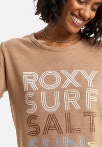 Glimlachende vrouw met een beige T-shirt met de tekst "ROXY SURF SALT SUN" en gelaagde gouden kettingen tegen een witte achtergrond.