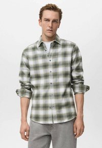 Camisa de franela a cuadros verde y blanca con frente de botones, mangas enrolladas y una textura suave. Llevada sobre una camiseta blanca, combinada con jeans grises.