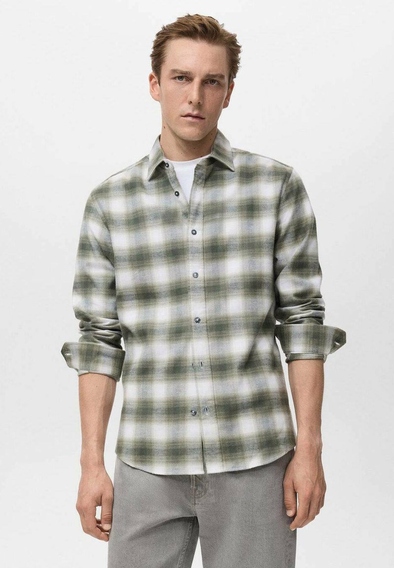 Camisa de franela a cuadros verde y blanca con frente de botones, mangas enrolladas y una textura suave. Llevada sobre una camiseta blanca, combinada con jeans grises.