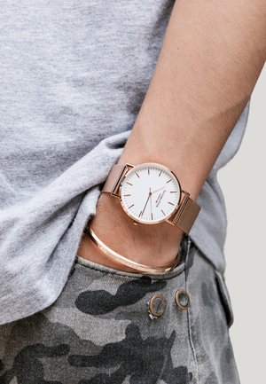 Sat od ružinog zlata s mrežastim remenom i bijelim brojčanikom, tankim oznakama sati, nošen na zapešću uz narukvicu od ružinog zlata, postavljen na sivu tkaninu i maskirne hlače.