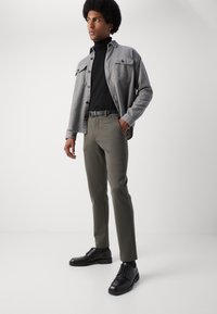 Chemise en flanelle grise superposée sur un col roulé noir, associée à un pantalon vert olive et des chaussures noires, avec un tissu texturé et une coupe droite.