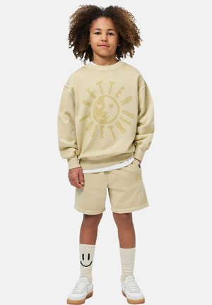 Beige sweatshirt met "Better Together"-opdruk, bijpassende korte broek, lichte sokken met een smiley en witte sneakers met beige zolen.