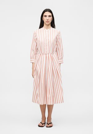 Frau mit langen dunklen Haaren, die ein weißes Midi-Kleid mit vertikalen roten Streifen, langen Ärmeln, vorne geknöpft und schwarze Flip-Flops trägt, steht gerade.