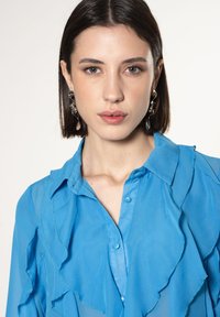 Lichtblauw semi-doorzichtig shirt met rimpeldetails en een knoopsluiting, met een klassieke kraag en een gladde textuur. Zilveren oorbellen zichtbaar.