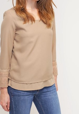 Frau trägt beige langärmlige Bluse mit Spitzenmanschetten und blaue Jeans, steht vor einem einfarbigen weißen Hintergrund.