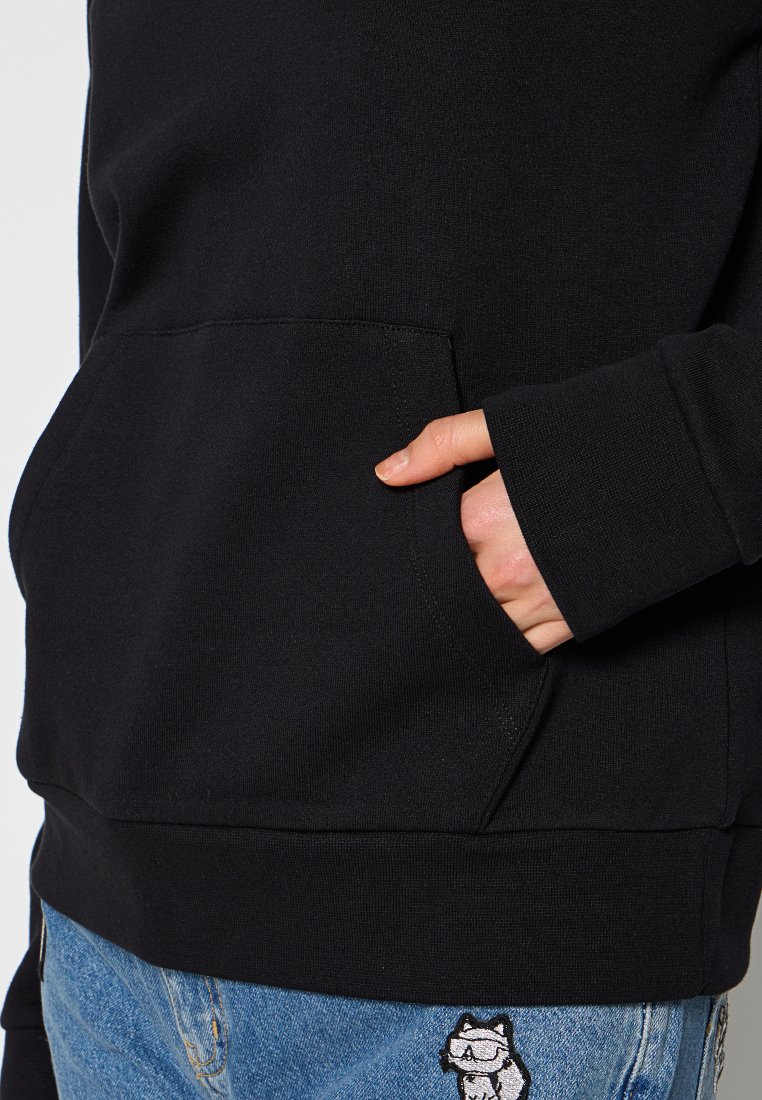 Felpa nera con finitura testurizzata, dotata di tasca frontale e polsini a coste; abbinata a pantaloni di jeans con una toppa a forma di gatto.