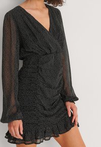 Vestido negro con lunares blancos, mangas largas transparentes, escote en V profundo, cinturilla fruncida y bajo con volantes acampanados. Tejido ligero.