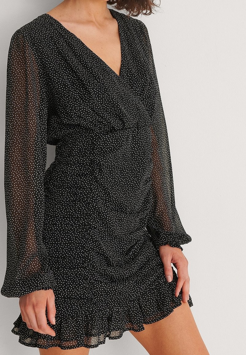 Vestido negro con lunares blancos, mangas largas transparentes, escote en V profundo, cinturilla fruncida y bajo con volantes acampanados. Tejido ligero.