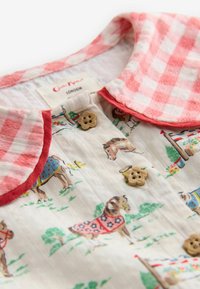 Balts bērnu apģērbs ar rozā rūtainu apkakli, zieda formā pogām un zirgu ilustrācijām, kas valkā segas un ir apkārt zaļajiem augiem.