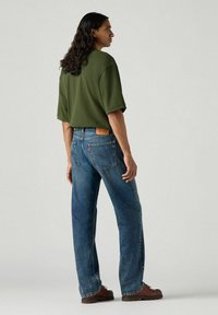 Green oversized short-sleeve t-shirt paired with blue straight-leg jeans. Brown footwear and visible denim stitching and hardware details.