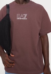 Brązowy bawełniany t-shirt z krótkimi rękawami, z białym logo "EA7 Emporio Armani". Czarna torba na ramię noszona na ramieniu.