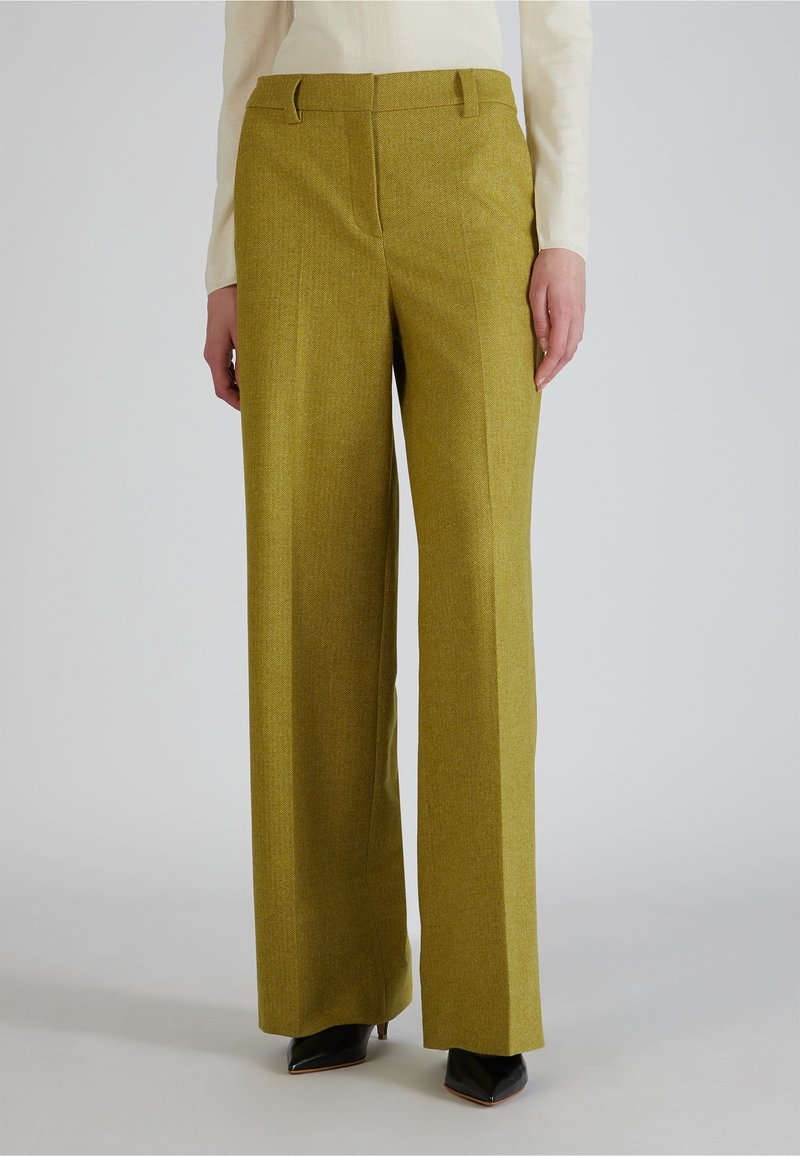 Vrouw in mosterdgele, hoge taille, wijde broek met voorplooi en zwarte puntige schoenen, staande tegen een effen achtergrond.