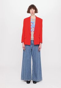 Red blazer with two front pockets over a light grey graphic t-shirt, paired with wide-leg blue denim pants featuring white stripes. Black shoes.