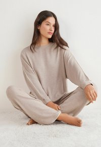 Beige loungedragt lavet af blød, tekstureret stof, der har en løs pasform, lange ærmer og brede bukser. Enkel design med en klassisk rund hals.