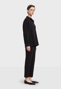 Zwarte colbert tuniek met een relaxte pasvorm, lange mouwen en knoopmanchetten, gecombineerd met zwarte toelopende broeken en platte schoenen.