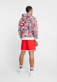 Hombre vestido con un sudadero con el logo de Coca-Cola, pantalones cortos rojos, calcetines blancos con rayas azul marino y rojas, y zapatillas blancas, de pie mirando hacia atrás.