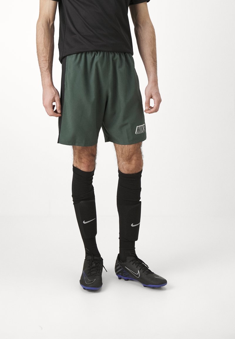 Homme portant un short Nike vert foncé, des chaussettes de football noires avec des logos Nike blancs, et des crampons Nike noirs, debout devant un fond blanc.
