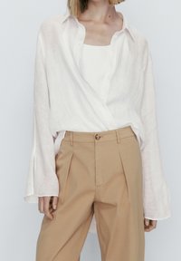 Chemise en lin léger blanche à manches longues, avec des boutons sur le devant, superposée à un débardeur beige ajusté et un pantalon beige taille haute.