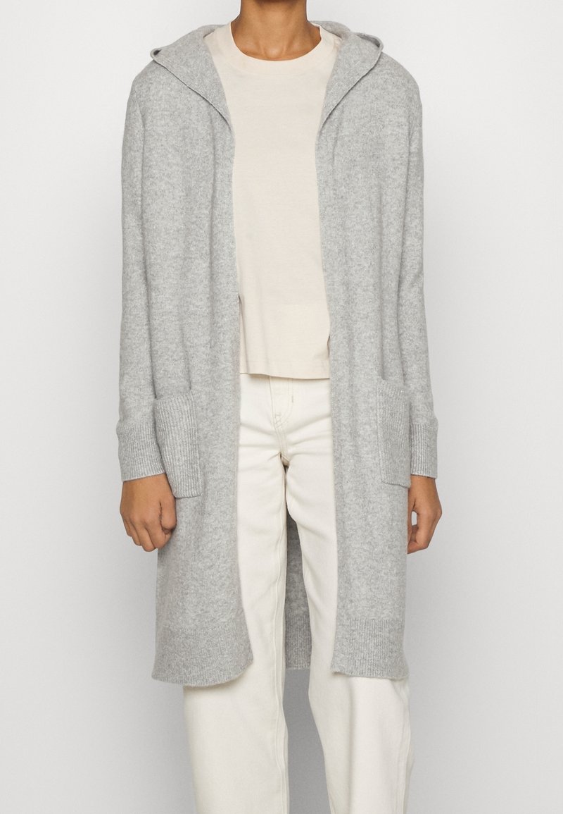 Personne portant un long cardigan à capuche gris clair sur une chemise beige et un pantalon blanc, debout devant un fond blanc uni.