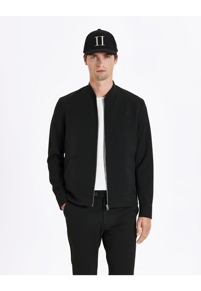 Veste bomber noire avec fermeture éclair à l'avant, col montant et poches latérales. Portée par-dessus un t-shirt blanc, associée à un pantalon noir et une casquette.