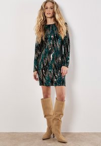 Sukienka z cekinami w ciemnozielonym, czarnym i srebrnym kolorze, z długimi rękawami i dopasowanym krojem. Połączona z beżowymi, zamszowymi kozakami do kolan.