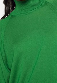 Maglione verde a collo alto con texture a costine, maniche lunghe e vestibilità rilassata. Dettagli delle cuciture visibili vicino al colletto e sulle spalle.