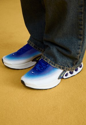 Osoba nosząca niebiesko-białe gradientowe sneakersy z czarnymi podeszwami i żółtymi akcentami, stojąca na dywanie koloru musztardowego.
