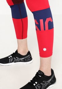 Czerwono-granatowe legginsy mają kolorowe bloki oraz widoczne szwy i logo. Połączone z czarnymi butami sportowymi z białymi akcentami.