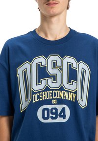 Modrá bavlnená tričko s veľkým bielym a žltým textom „DC Shoe Company“ a „094“, s kulatým výstrihom a krátkymi rukávmi.
