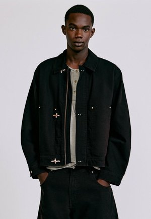 Jeune homme à la peau foncée portant une veste noire par-dessus une chemise rayée, debout les mains dans les poches devant un fond clair uni.
