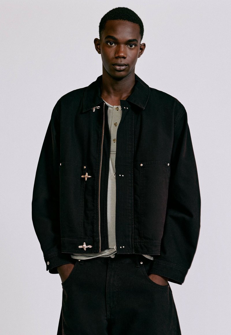 Jeune homme à la peau foncée portant une veste noire par-dessus une chemise rayée, debout les mains dans les poches devant un fond clair uni.