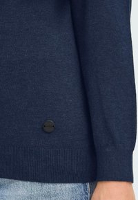 Marinblå stickad tröja med ribbad mudd och fåll, med en rund svart logotagg på nedre vänstra sidan. Mjuk textur, figursydd design.