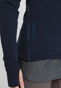 Tummansininen fleecejakku, jossa on vetoketjullinen tasku, ommeltuja yksityiskohtia ja turkoosi logo sivussa, sekä sileä ja pehmeä pinta.
