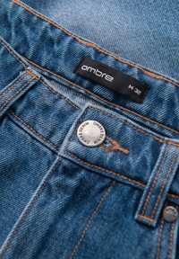 Blå denimjeans med orange syninger, med et sort "ombre" mærke og en sølvknap indgraveret med "OMBRE DENIM."