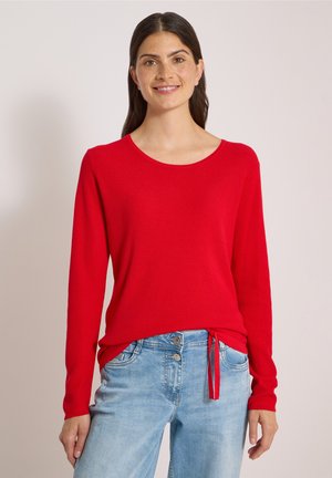 Donna sorridente con lunghi capelli scuri, che indossa una maglia rossa a maniche lunghe e jeans azzurri, in piedi davanti a uno sfondo chiaro.