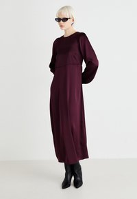 Burgundin pitkähihaineen mekko, joka on valmistettu sileästä kankaasta, sisältää löysän istuvuuden ja korkean kaula-aukon. Päällä mustat kärkitossut ja aurinkolasit.