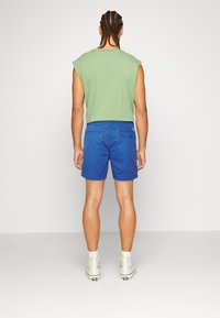 Camiseta verde sin mangas, pantalones cortos azules y zapatillas deportivas altas blancas. El atuendo tiene un ajuste casual con una textura suave y un diseño minimalista.
