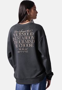Sudadera gris oscuro con un corte relajado, puños y dobladillo acanalados, y gran texto beige impreso en la parte posterior. Textura de tela suave y lisa.