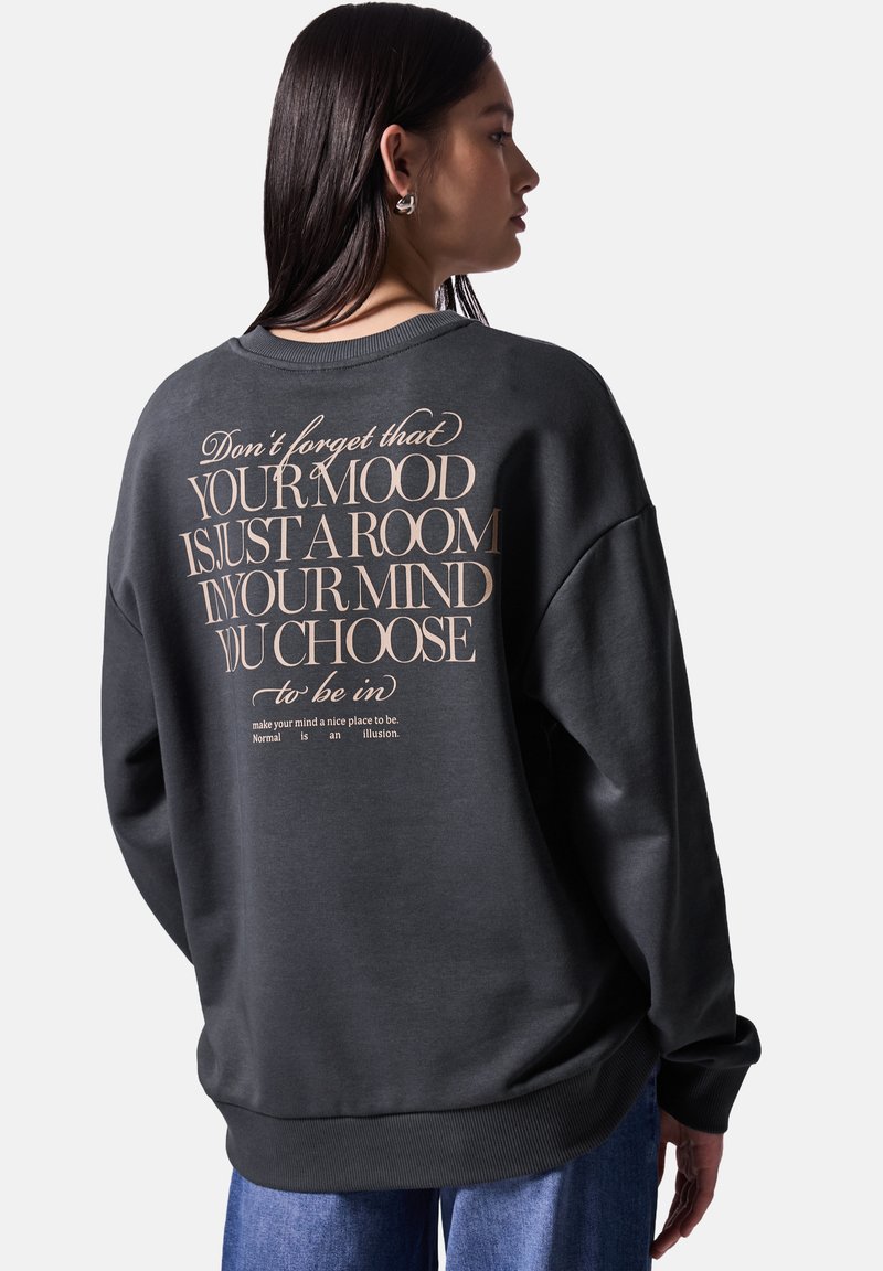 Sudadera gris oscuro con un corte relajado, puños y dobladillo acanalados, y gran texto beige impreso en la parte posterior. Textura de tela suave y lisa.