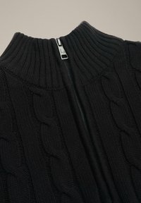 Czarny sweter z dzianiny warkoczowej z wysokim kołnierzem i srebrnym zamkiem błyskawicznym z przodu. Teksturowany wzór i prążkowane detale podkreślają design.