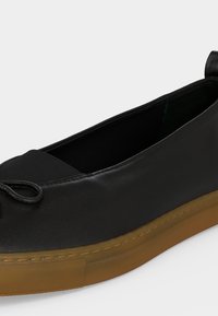 Μαύρο δερμάτινο επίπεδο παπούτσι με καφέ καουτσούκ σόλα και διακοσμητικό φιόγκο στο δάχτυλο, πλάγια όψη που δείχνει το εσωτερικό και το πλάι.