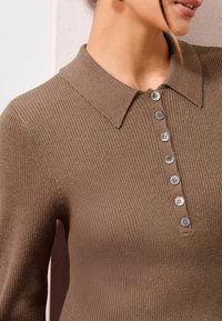 Polo en maille côtelée marron avec col et patte de boutonnage comportant six boutons gris clair. Gros plan sur le haut du vêtement.