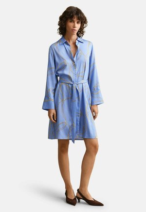 Vrouw draagt een koningsblauwe shirtjurk tot op de knie met gouden kettingpatronen, een gestrikte riem in de taille, lange mouwen en bruine slingbacks met hak.