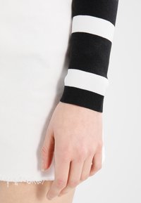 Schwarz-weiße gestreifte Strickärmel mit einer enganliegenden Textur, kombiniert mit einem weißen Denim-Kleidungsstück mit fransigem Saum. Hand im Bild.